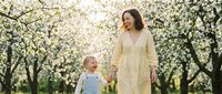 Happy mother and toddler son walking hand in hand through blooming spring orchard with white flowers creating magical family bonding moment outdoors.