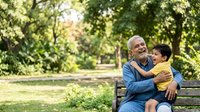 Mature man with gray hair and glasses, South Asian ethnicity, sitting on park bench with young boy, embracing and smiling together outdoors among green trees and foliage for family bonding concepts.