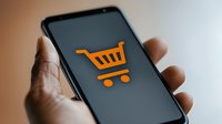 Orange shopping cart icon displayed on smartphone screen held in hand against blurred background, representing mobile commerce and online shopping application.