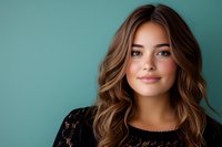 Young Hispanic woman with wavy brown hair and natural makeup smiling confidently against turquoise background wearing black lace top, studio portrait.