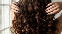 Woman with voluminous curly brown hair touching her locks with manicured hands in bright natural light for beauty and haircare concepts.