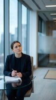 Professional businesswoman in dark suit sitting at glass conference table in modern office with city view looking thoughtful during corporate meeting.