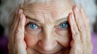 Senior woman with bright blue eyes holding her face in her hands, expressing emotion with a gentle smile, showcasing aging and wisdom.