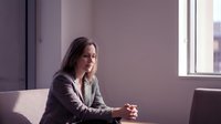 Thoughtful Caucasian businesswoman sitting in modern office, hands clasped, looking down in focused contemplation near bright window.