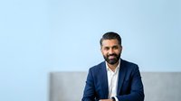 Young Middle Eastern male professional in navy blazer and white shirt sitting on gray couch against light blue background. Corporate headshot for business, leadership, and corporate communications use.