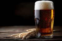 Cold glass of dark craft beer with foam head and wheat ears on rustic wooden table against black background, dramatic lighting highlights amber liquid.