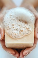 Natural soap bar with foam bubbles held in feminine hands against white background, close up view showing texture and lather for skincare and hygiene concept.