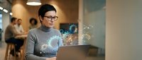 Asian businesswoman with glasses using laptop with digital holographic interface icons in modern office workspace for technology presentations.