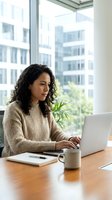 Young woman of Latin American descent working on laptop at wooden desk in modern office with city skyline view, wearing beige sweater, suitable for business and corporate concepts.