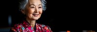 Smiling senior Asian woman with gray hair wearing floral blouse against dark background, expressing joy and confidence in portrait style photography.