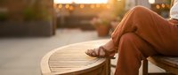Woman in brown pants relaxing on outdoor wooden bench during golden hour sunset with warm bokeh lights creating peaceful urban atmosphere for lifestyle content.
