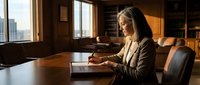 Middle-aged Caucasian female professional in business attire working at wooden desk in upscale office with city skyline view, suitable for corporate, business, and workplace concepts.