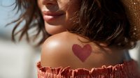Young woman with heart-shaped temporary tattoo on shoulder wearing off-shoulder coral top at beach, sunlight highlighting skin tone and wavy hair.