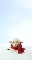 Cute sheep toy wearing red knitted scarf sitting on snow with blue sky background, suitable for holiday greeting cards and winter seasonal designs.