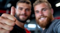 Two smiling bearded men taking selfie and showing thumbs up gesture at gym, expressing positivity and achievement after workout session.