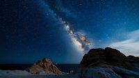 Majestic Milky Way galaxy arches over dramatic rocky coastal cliffs at night, with starry sky and ethereal blue glow creating mystical natural landscape.