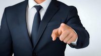 Businessman in navy blue suit pointing finger forward against light background, conveying leadership and direct communication in professional setting.