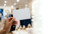 Hand holding blank white card in modern office or retail environment with blurred background and warm lighting for business branding.