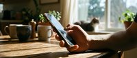 Caucasian man using smartphone at wooden kitchen table in warm morning sunlight, with coffee mugs and cat in background.