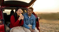 Young Asian woman and Caucasian man enjoying coffee break during road trip, sitting in car trunk at sunset. Couple sharing romantic moment outdoors.