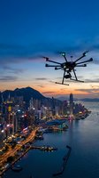 Professional drone flying over illuminated city skyline at sunset with harbor view and mountain backdrop for aerial photography and videography projects.