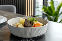 Tender braised beef with colorful root vegetables and potatoes in modern white ceramic bowl, garnished with fresh herbs and lemon wedge. Restaurant quality meal.