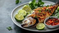 Grilled whole fish with lime slices and spicy sauce on plate. Fresh herbs and citrus garnish create appetizing seafood meal for restaurant menu or cooking blog.