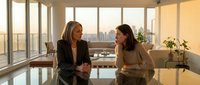 Two professional businesswomen having meeting in modern office with city skyline view during golden hour sunset light through floor-to-ceiling windows.