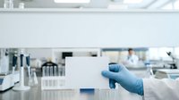 Scientist in blue latex glove holding blank white card in modern laboratory with equipment and blurred background, suitable for medical research, healthcare, and scientific communication projects.