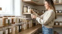 Young woman organizing glass jars with grains and spices on wooden shelves in modern zero waste kitchen pantry for sustainable living lifestyle.