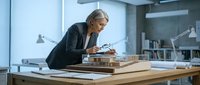Senior male architect reviewing wooden building models in modern office workspace with blueprints and design materials for construction planning.