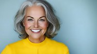 Confident mature woman with natural gray hair wearing yellow sweater smiling at camera against blue background, representing vitality and positive aging.