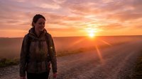 Young adult male hiker with backpack standing on mountain trail at golden hour sunrise, watching dramatic orange and yellow sky over valley landscape for travel and adventure concepts.