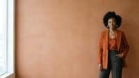 Young woman with dark curly hair and medium skin tone wearing burnt orange blazer and olive pants standing against warm terracotta wall with natural light from window.