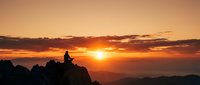 Person silhouette meditating on mountain peak during golden sunset with dramatic orange sky and layered mountain ranges creating peaceful spiritual moment.