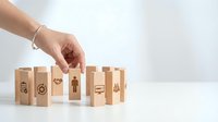Female hand arranging wooden blocks with business icons on white background, symbolizing teamwork, strategy, and corporate management concepts.