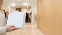 Woman holding blank white tablet in modern retail clothing store with warm lighting and wooden interior design.