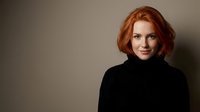 Portrait of confident young Caucasian woman with red hair wearing black turtleneck sweater against neutral background for professional headshot.