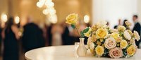 Elegant wedding reception table with yellow and cream rose bouquet in soft candlelight with blurred guests celebrating in luxurious venue background.