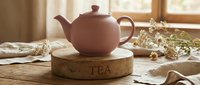 Matte pink ceramic teapot on a rustic wooden stand with TEA inscription, surrounded by dried flowers in a cozy kitchen setting.