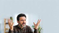 Middle-aged man with dark curly hair and beard, appearing frustrated or confused, gesturing with both hands raised. Wearing brown sweater in bright office setting with plants.