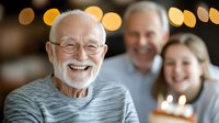 Senior Caucasian man with white beard and glasses laughing at birthday celebration with family, lit candles and bokeh lights creating warm atmosphere.