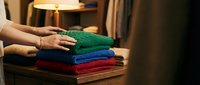 Woman organizing colorful knitted sweaters stack on wooden table in cozy home interior with warm lighting for seasonal wardrobe preparation.