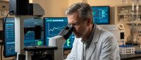 Middle-aged Caucasian male scientist in white lab coat examining samples through microscope in modern research laboratory with digital displays and equipment.