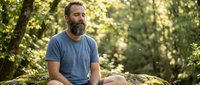 Middle-aged man with full beard and dark hair, Caucasian ethnicity, wearing casual blue t-shirt, sitting outdoors on rock in forest setting with natural sunlight filtering through trees.