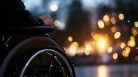 Person in wheelchair at dusk against blurred city lights background creating atmospheric mood with bokeh effect, representing disability awareness and inclusion.