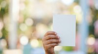 Young child with medium skin tone holding blank white card outdoors with blurred green and colorful background, suitable for advertising, marketing campaigns, and promotional messaging.