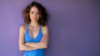 Young woman with curly hair wearing blue tank top standing with arms crossed against purple background, confident pose for lifestyle or fashion content.