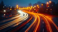 Long exposure night photography of curved highway with glowing light trails from moving vehicles creating dynamic streaks against dark blue evening sky with bokeh effects.