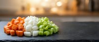 Fresh diced vegetables including orange carrots, white onions and green celery arranged on dark slate board in modern kitchen for cooking preparation.